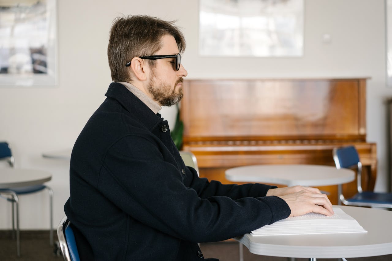 hero-img-01 A visually impaired man reading Braille at a table in a warmly lit room with a piano in the background.